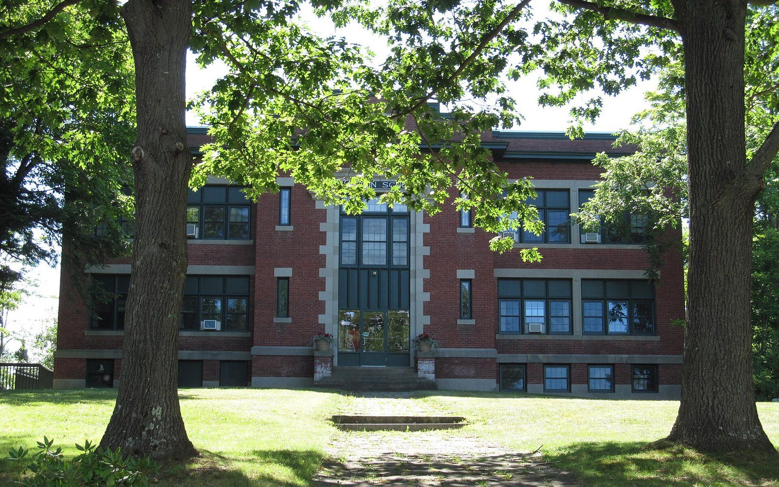 A brick school building with tall arched windows framed by two large trees